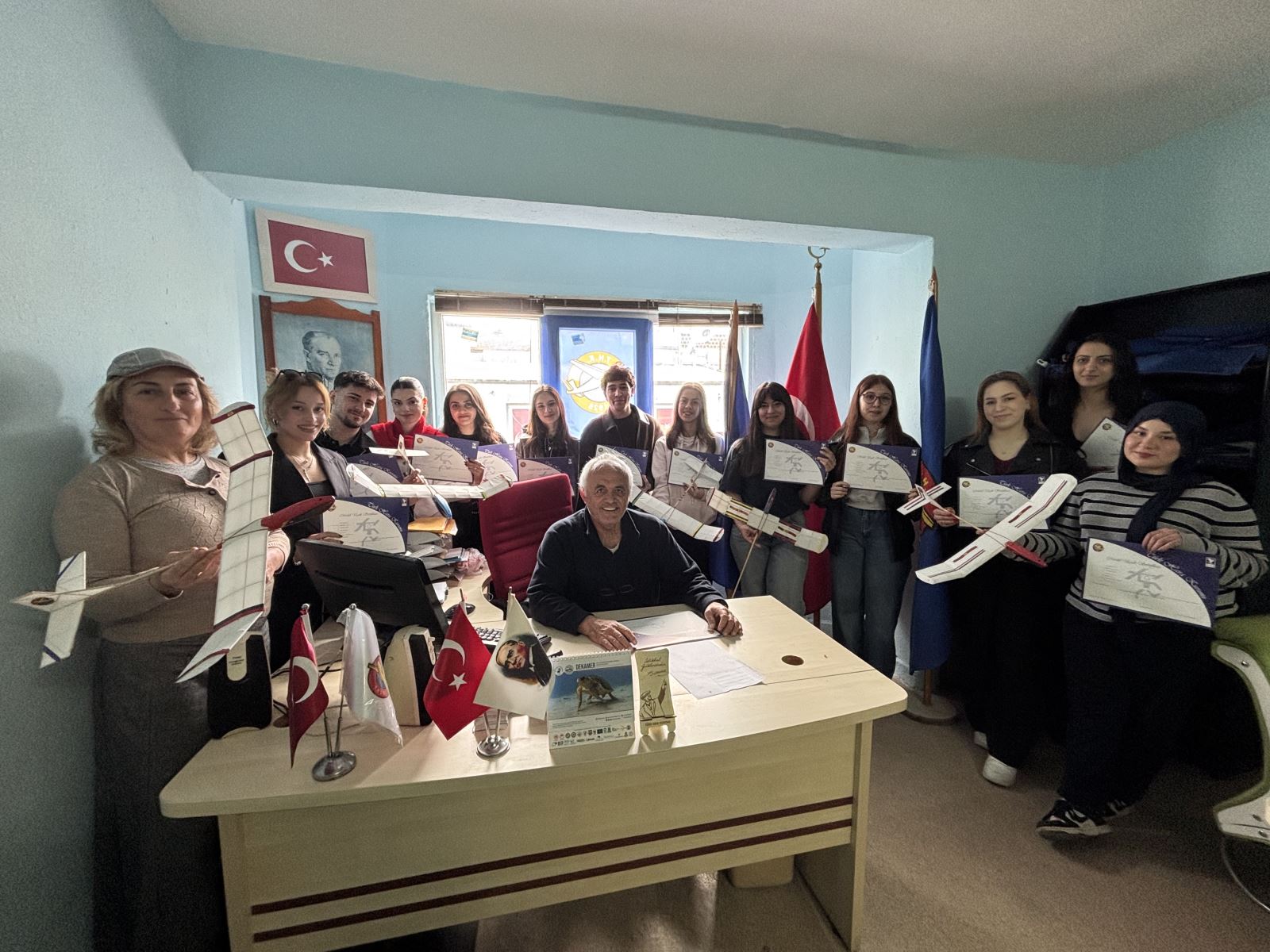 Atölye katılımcıları ve eğitmenleri bir masa etrafında toplanmış, üzerinde "Türk Hava Kurumu Ortaca Şubesi" yazılı bir bayrağın arkasında duruyor. Masanın üzerinde model uçak parçaları ve malzemeleri var.